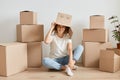 Indoor shot of young adult Caucasian woman wearing funny cardboard box on head sitting on floor with crossed legs at new home,