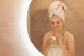 Indoor shot of young adult female taking care of her skin, posing in bathroom in front of mirror with cosmetic cream in hands, Royalty Free Stock Photo