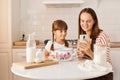 Indoor shot of young adult woman cooking with her daughter in the kitchen, browsing internet for finding recipe or broadcasting