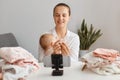 Indoor shot of young adult woman posing with infant daughter in front of tripod with smartphone, filming video for her vlog,