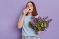 Indoor shot of young attractive sick female wearing blue t shirt standing isolated over purple background and holding lilac