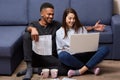 Indoor shot of young beautiful couple, sitting on floor, using their laptop, having pleasant communication, smiling sincerely, Royalty Free Stock Photo