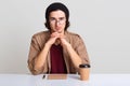 Indoor shot of young man keeps hands under chin, looks with self confident facial expression, wears glasses, cap, shirt and jacket Royalty Free Stock Photo