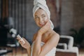 Indoor shot of young smiling woman applies moisturizer cream on face, takes care of her skin and complexion, puts lotions, has Royalty Free Stock Photo