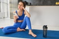 Indoor shot of young sportswoman holding smartphone, sits with fitness water bottle, uses sports app to workout, do yoga Royalty Free Stock Photo