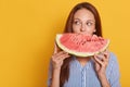 Indoor shot of young woman biting big slice of watermelon while looking aside, wearing striped shirt, posing isolated over yellow Royalty Free Stock Photo