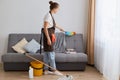 Indoor shot of young woman cleaning her home, wearing jeans and brown apron, housewife doing her household chores, standing near Royalty Free Stock Photo