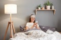 Indoor shot of young woman wearing sleeping mask sitting in bed using mobile cell phone, typing sms message, resting at home, Royalty Free Stock Photo