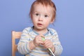 Indoor studio picture of curious concentrated infant sitting on chair, looking up, holding toy in both hands, playing alone,
