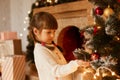 Indoor studio shot of charming girl wearing white sweater and having pigtails, decorating Christmas tree, standing near fireplace Royalty Free Stock Photo