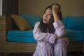 Young beautiful sad and depressed Asian Chinese woman in pajamas at home sitting by living room couch feeling unhappy and confused