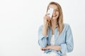 Indor shot of satisfied carefree Caucasian blond woman in stylish clothes, holding smartphone over eye and smiling