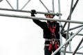 An industrial climber wearing a helmet and a protective belt is tied with a rope to a support at a high altitude. A specialist