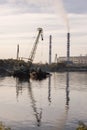 Industrial landscape with cranes and pipes. The plant works near the river. Crane on a barge loads cargo. Industrial area