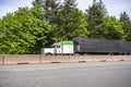 Classic big rig green and white semi truck with long cab transporting cargo in covered black semi trailer running on the divided Royalty Free Stock Photo