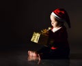 Infant baby boy in red santa claus costume and cap is occupied with a golden gift box, holding, looking how to untie