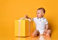 Infant baby boy toddler is sitting at big yellow gift box with ribbon and bow, going to untie on yellow background