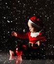 Infant baby boy toddler is sitting in santa claus costume, cap under the snow playing with big red christmas tree ball Royalty Free Stock Photo