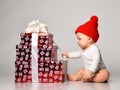 Infant baby boy toddler in white bodysuit and red hat is occupied with new year christmas gift boxes holding ribbon