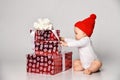 Infant baby boy toddler in white bodysuit and red hat is sitting at gift boxes unpacking preparing on white