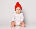 Infant baby boy toddler in white bodysuit and red warm hat with pompon is sitting looking directly at camera on white