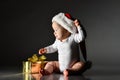 Infant baby boy toddler in white bodysuit and santa claus hat is sitting with golden new year christmas gift box on dark