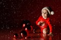 Infant baby girl in red bodysuit and Santa hat sits at Christmas New Year tree balls and looks at corner under snow Royalty Free Stock Photo