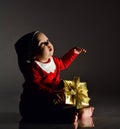 Infant boy toddler in santa claus costume is sitting with new year gift box present holding hand up, slapping, looking Royalty Free Stock Photo