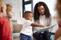 Infant school boy pointing in a book held by the female teacher, sitting with kids on chairs in the classroom, close up Royalty Free Stock Photo