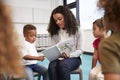 Infant school boy pointing in a book held by the female teacher, sitting with kids in a circle on chairs in the classroom, close u Royalty Free Stock Photo