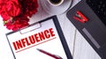 INFLUENCE is written in red on a white notepad near a laptop, coffee, red roses and a pen