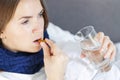 Influenza treatment or coronavirus . Flu-sick woman, wrapped in warm scarf and blanket , taking pills holding glass of water Royalty Free Stock Photo