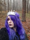 An informal goth girl with purple hair and a silvery crown with stars is walking in a forest