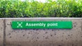 Informative green assembly point sign with people icon graphic on wall with hedge outside in public place in England. Royalty Free Stock Photo