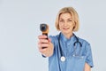 With infrared thermometer. Middle-aged professional female doctor in uniform and with stethoscope