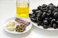 Ingredients for tapenade: pitted black olives on white plate, olive oil in glass bottle, three cloves of garlic, some anchovy