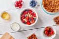 Ingredirnts for preparing healthy organic breakfast - granola, berries honey, milk, banana in white bowl on white table