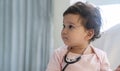 Innocence smile toddler girl sitting on the white bed with stethoscope while looking something at home. Mixed race little doctor Royalty Free Stock Photo