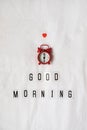 Inscription Good morning, red analog clock, small heart on white rumpled sheets. Top view, flat lay. Vertical. Concept of rest,