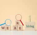 The inscription we are hiring and a wooden figurine on the podium with a plastic magnifying glass on a beige background. Employee