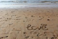 Inscription of `Love` and a symbol of heart in the sand. Footprints in the sand and sea waves at sunset. Royalty Free Stock Photo