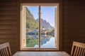 Inside a rorbu, looking through an open window, the calm Norwegian fjord mirrors quaint houses and a rugged cliff