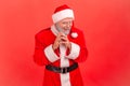 Insidious elderly man with gray beard wearing santa claus costume looking at camera with cunning tricky face and smirk, planning