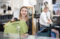 Girl stopped near showcase with outerwear, holds blouse hanger in hands and examines product