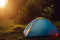 Inspiration camping place with blue tent during sunrise. Beautiful sunbeams through summer forest. Amazing scene of campsite in Royalty Free Stock Photo