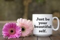 Inspirational message on a white mug - Just be your beautiful self. Message on coffee cup with soft pink flowers on wooden table. Royalty Free Stock Photo