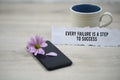 Inspirational quote - Every failure is a step to success. With morning cup of coffee and purple daisy flower on black phone.