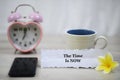 Inspirational quote - The time is now. With blurry white background of a cup of morning coffee, black phone, pink alarm table Royalty Free Stock Photo