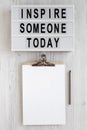 `Inspire someone today` words, clipboard with blank sheet of paper on a lightbox on a white wooden background, top view. Overhea