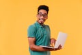 Inspired african student in glasses holding white laptop on yellow background. Studio shot of caref